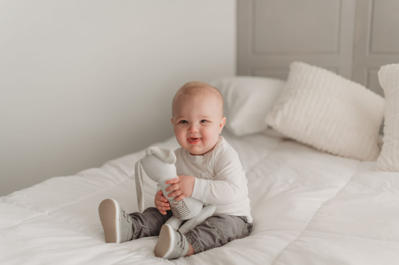 One year old little boy sitting on a bed with white sheets and white pillows, wearing a white shirt, gray pants, and gray shoes, smiling, and holding a stuffed bunny