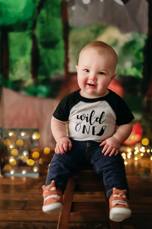 One year old little boy wearing jeans, a white and black shirt that writes "wild one", with blurred out lights and a forest background