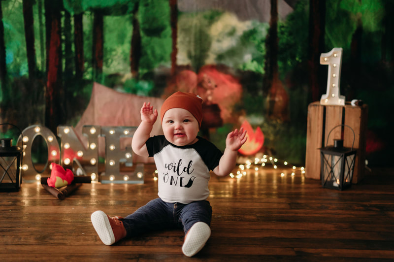 One year old little boy wearing jeans, a white and black shirt that writes "wild one", a fox hat, fox shoes, with blurred out lights and a forest background