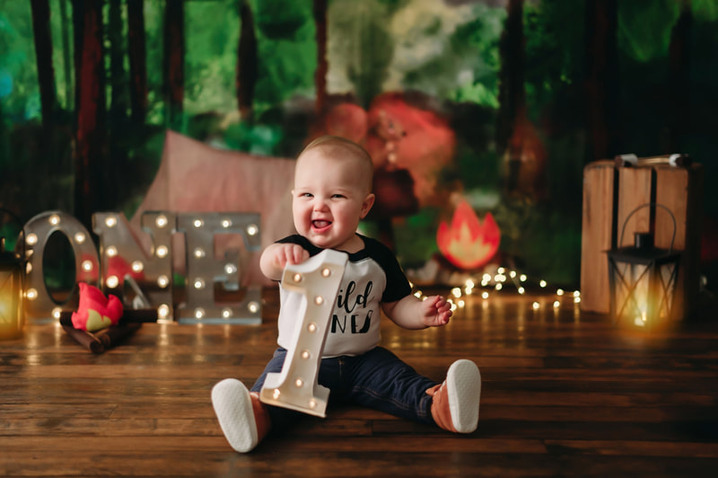 One year old little boy holding a lite up number one, wearing jeans, a white and black shirt that writes "wild one", with blurred out lights and a forest background