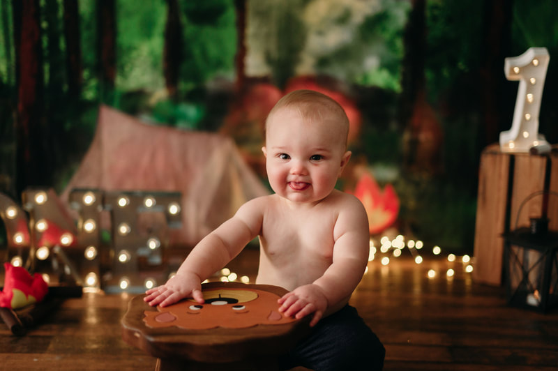 One year old little boy wearing jeans, with blurred out lights and a forest background. He is playing on a stole that has a bears face on it.