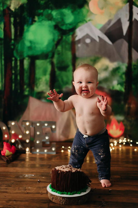 One year old little boy wearing jeans, with blurred out lights and a forest background. He has cake all over his face, hands, and pants.