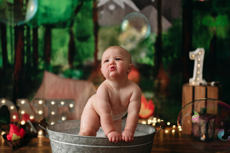 One year old little boy naked in a small bucket tub taking a bath, with blurred out lights and a forest background.