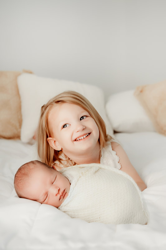 baby boy on a white bed with his older sister swaddled in a cream blanket while sister holds him