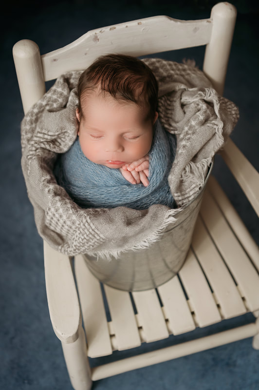 sleeping baby boy in a silver bucket on a white chair swaddled in a blue blanket