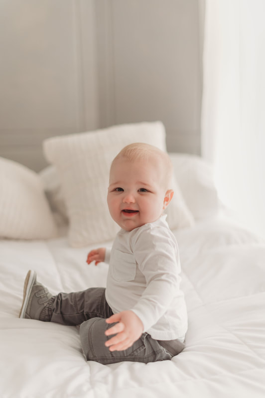 One year old little boy sitting on a bed with white sheets and white pillows, wearing a white shirt, gray pants, and gray shoes, smiling.