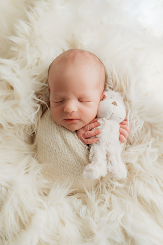 baby boy sleeping on a white fur blanket, swaddled in a cream rap holding a cream colored teddy bear