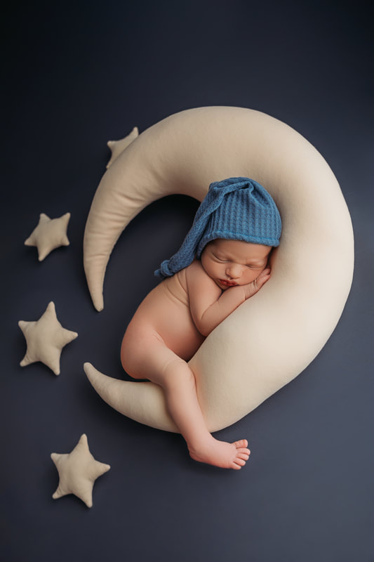 baby boy cuddling a light yellow moon with small stars around him on a dark blue blanket wearing a blue hat