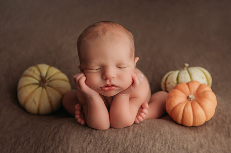 baby boy on a brown blanket surrounded by 3 small pumpkins, one orange and two white with green