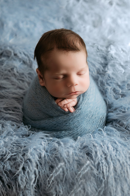 sleeping baby boy swaddling in a blue blanket sat up on a blue furry blanket