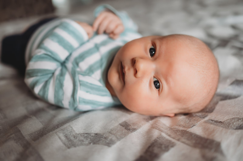Newborn baby boy laying on a bed looking at the camera. He is wearing a blue and white stripped shirt.